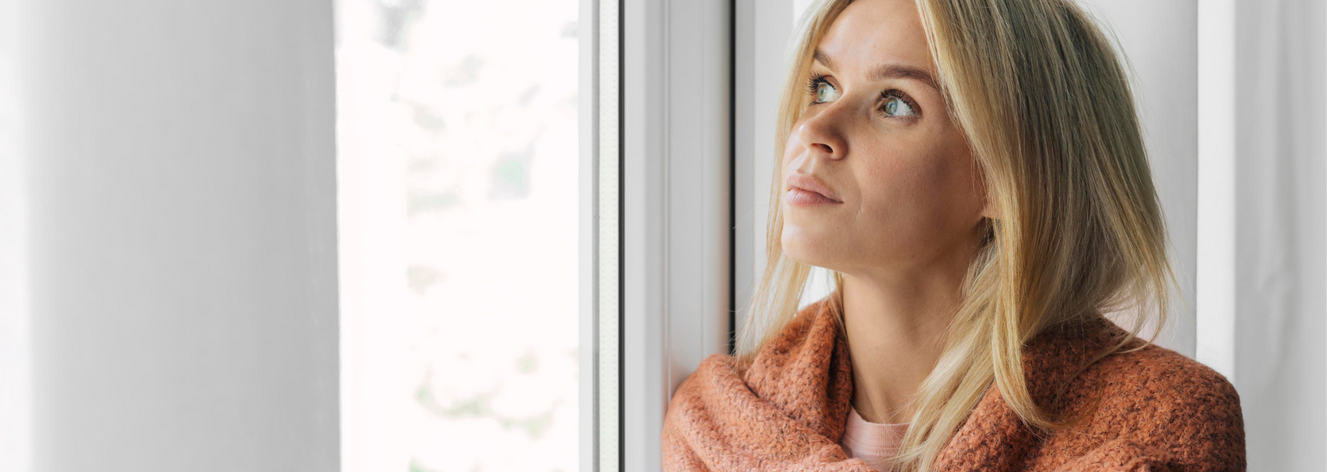 Blonde woman in an orange sweater looking out a bright window