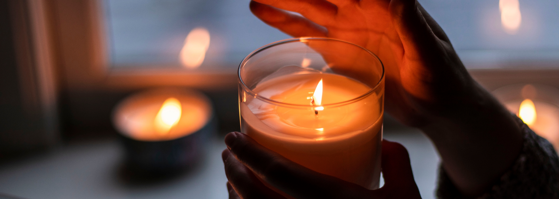 Hands holding a lit candle beside a blurred candle in warm orange light