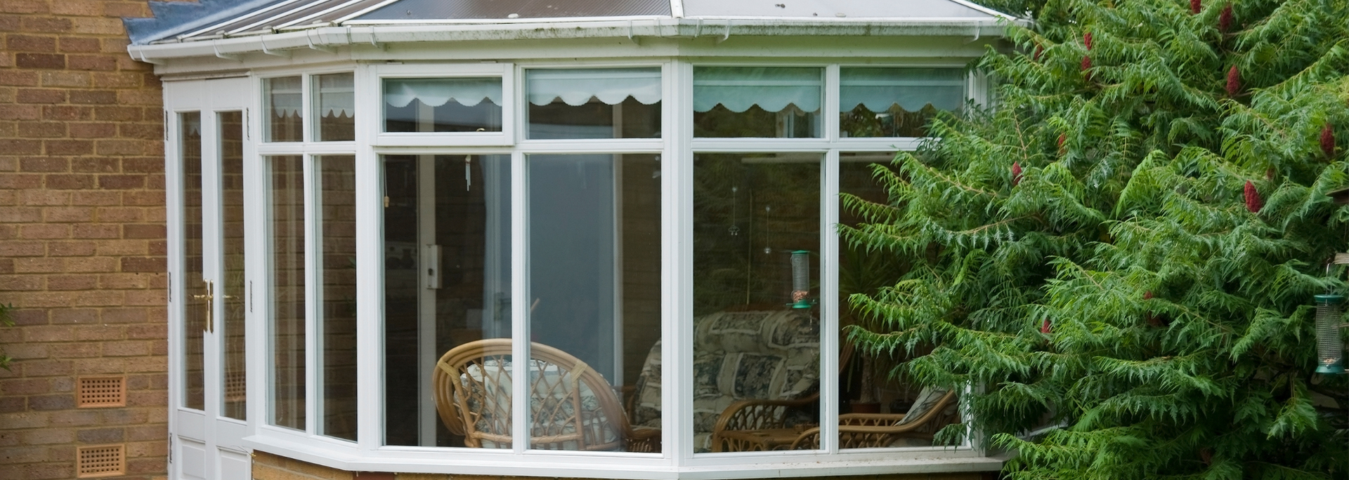 White sunroom with large windows attached to a brick house, partly hidden by green trees.