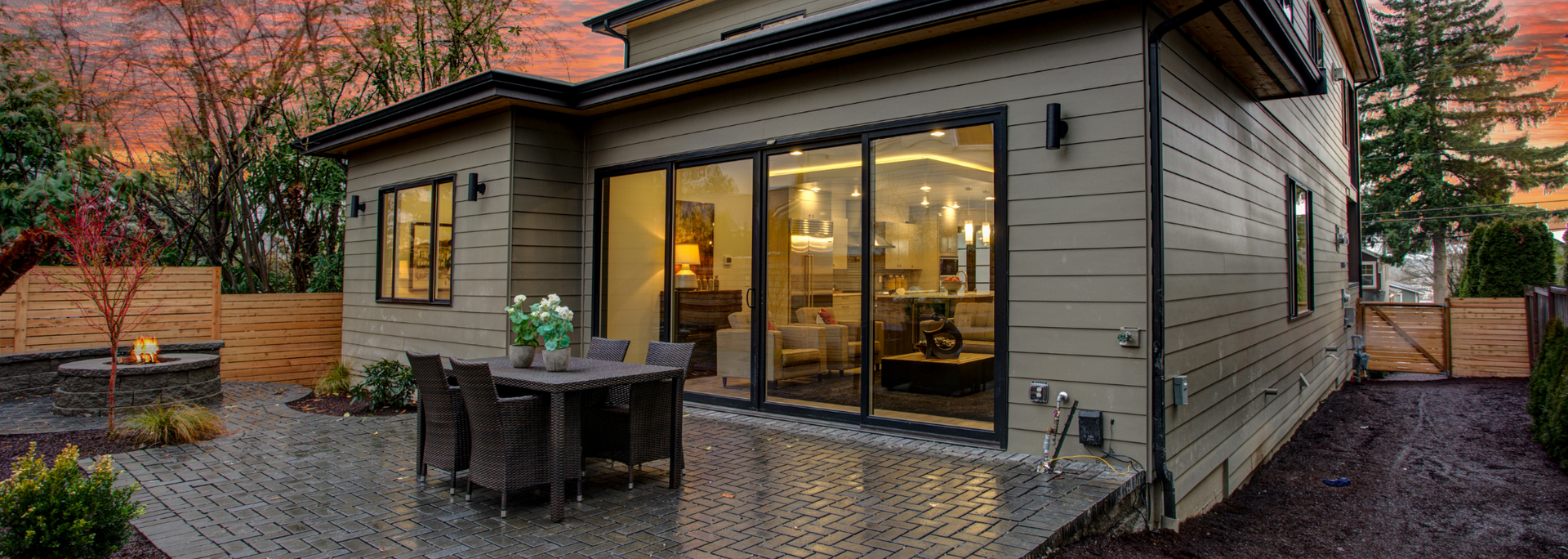 Modern house patio with sliding glass doors, outdoor dining set, and warm interior lights at dusk
