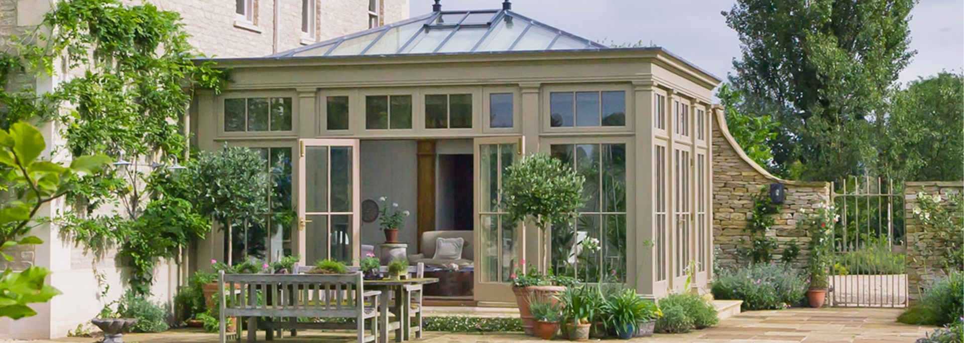 Small glass garden pavilion surrounded by greenery, with a table and chairs in front