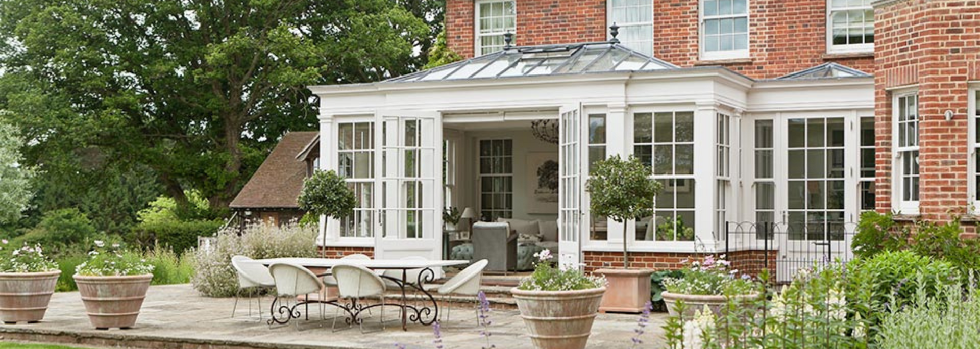 White sunroom patio with wicker furniture, potted plants, and brick house exterior surrounded by greenery