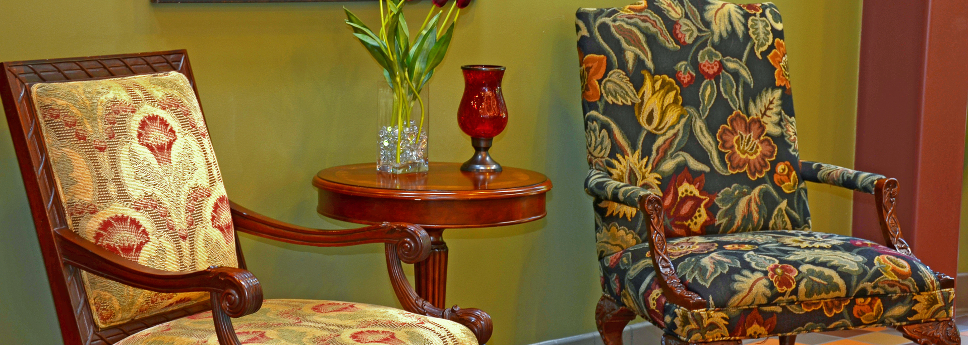 Two patterned armchairs beside a small round table with flowers and a red vase on a green wall background