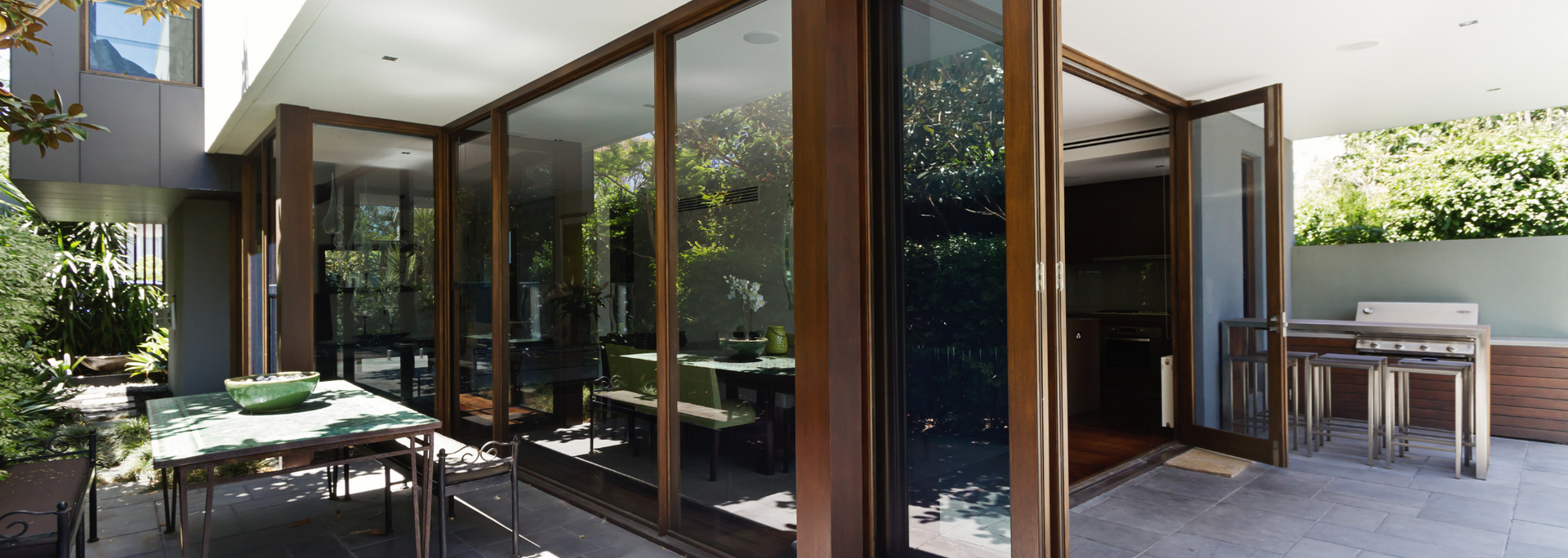 Modern patio with glass walls, outdoor seating, and lush greenery beside a house