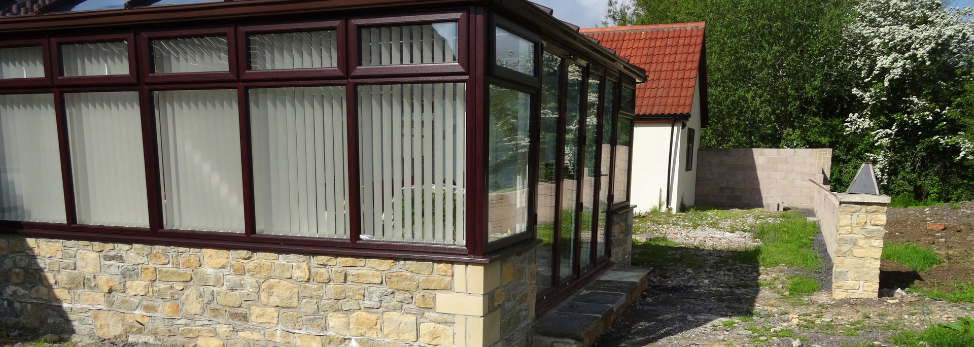 Stone-and-glass sunroom beside a garden path, with red roof and closed white curtains.