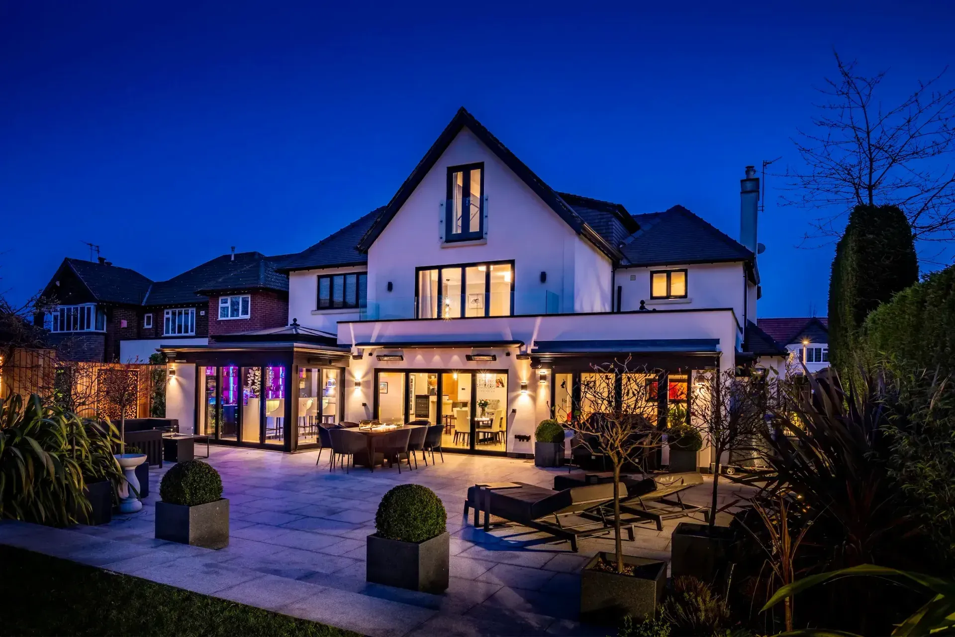 Illuminated modern house exterior at dusk with patio seating and warm indoor lights
