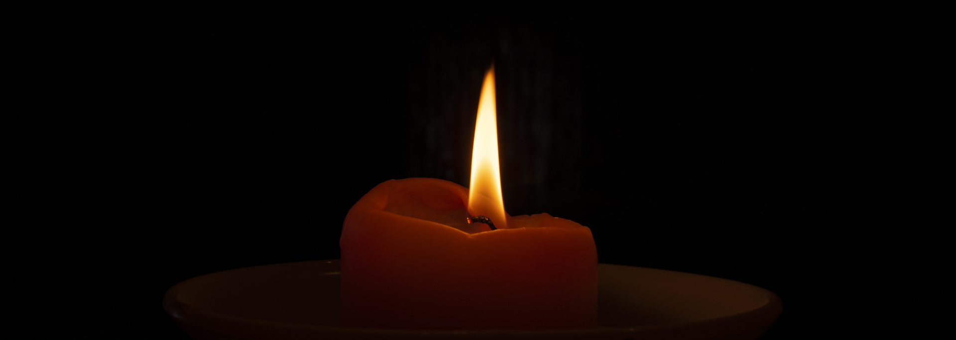 A single lit candle glowing in the dark on a saucer, with a warm orange flame.