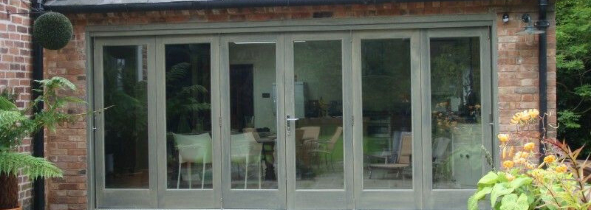 Front of a brick house with large gray-framed glass doors and windows, flanked by greenery.