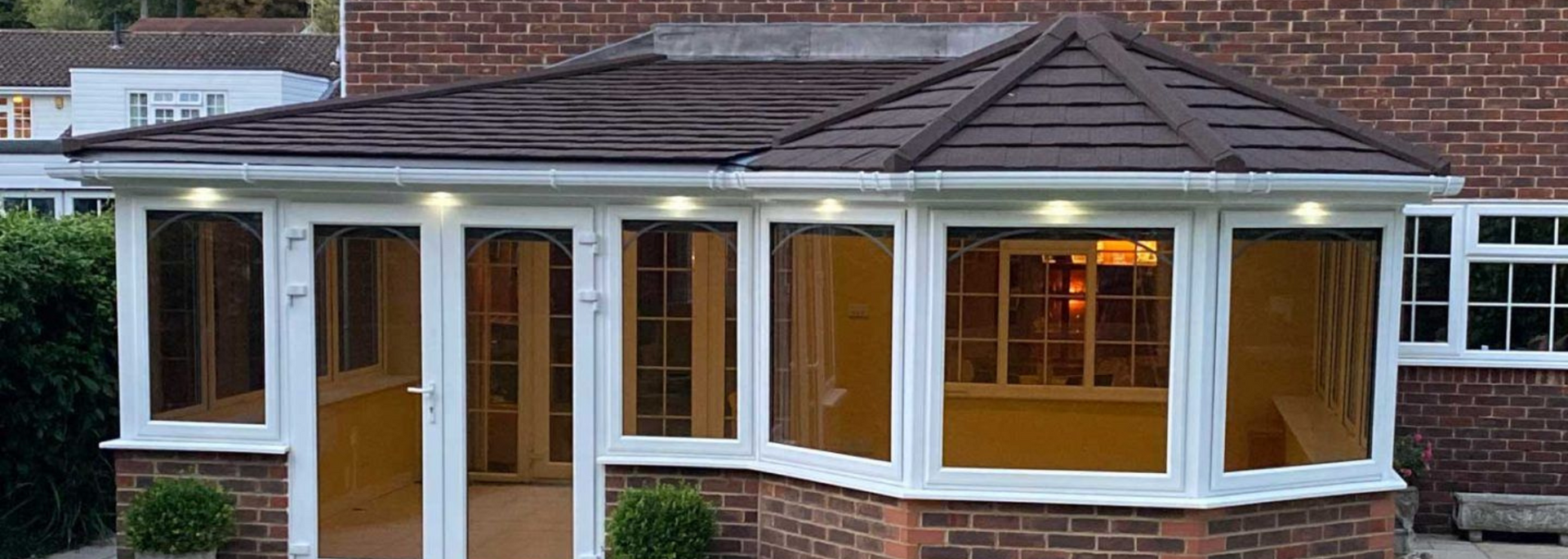 Brick house front with white-framed bay windows and a brown tiled roof at dusk