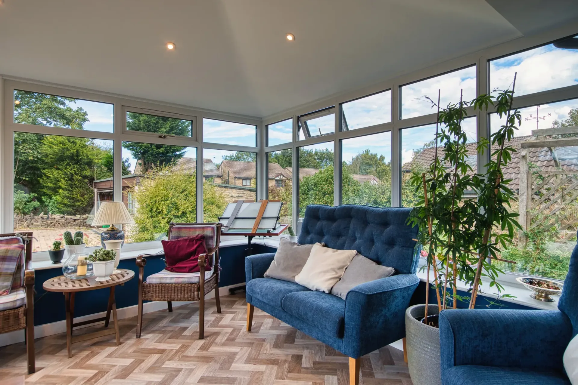 Bright sunroom with blue sofas, round table, potted plants, and large windows overlooking a garden.