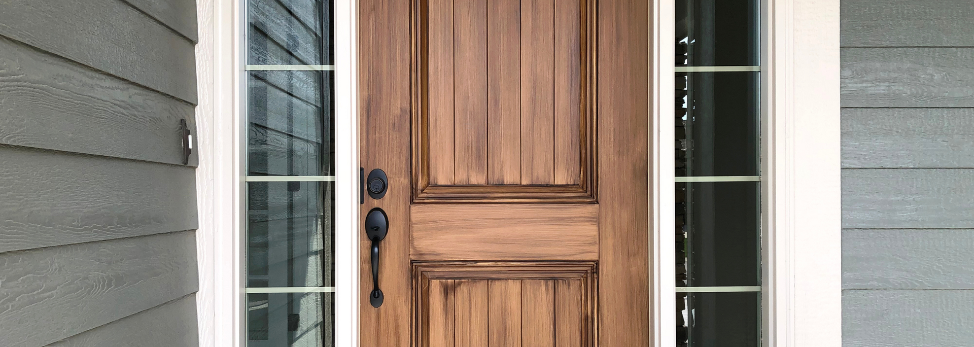 Wooden front door with black handles, flanked by narrow glass sidelights on a gray house exterior