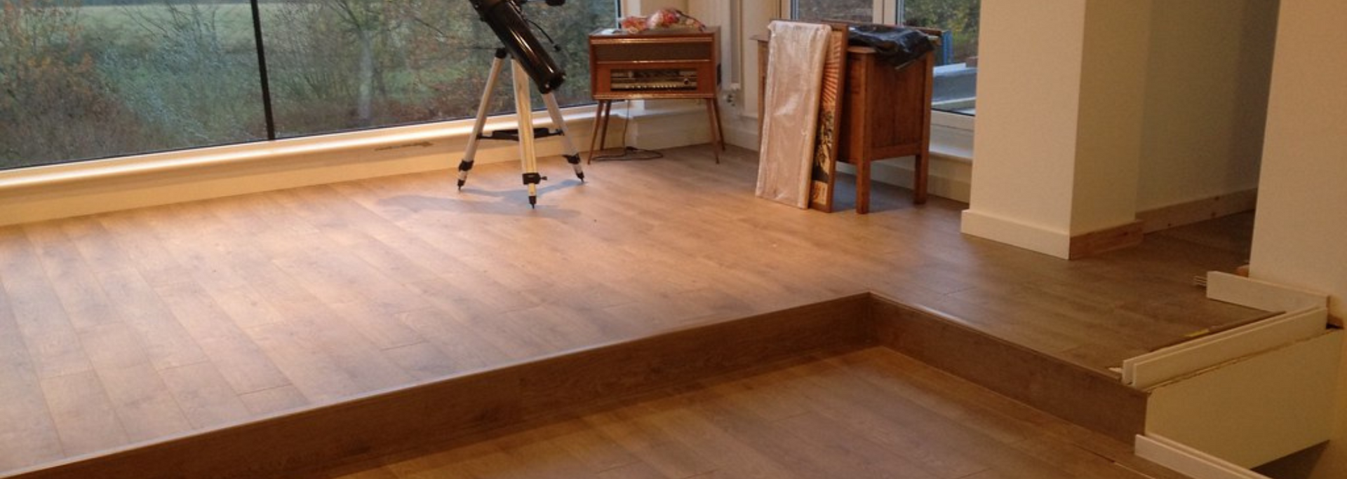 Empty wooden room with a raised platform, large windows, and a tripod near the back wall