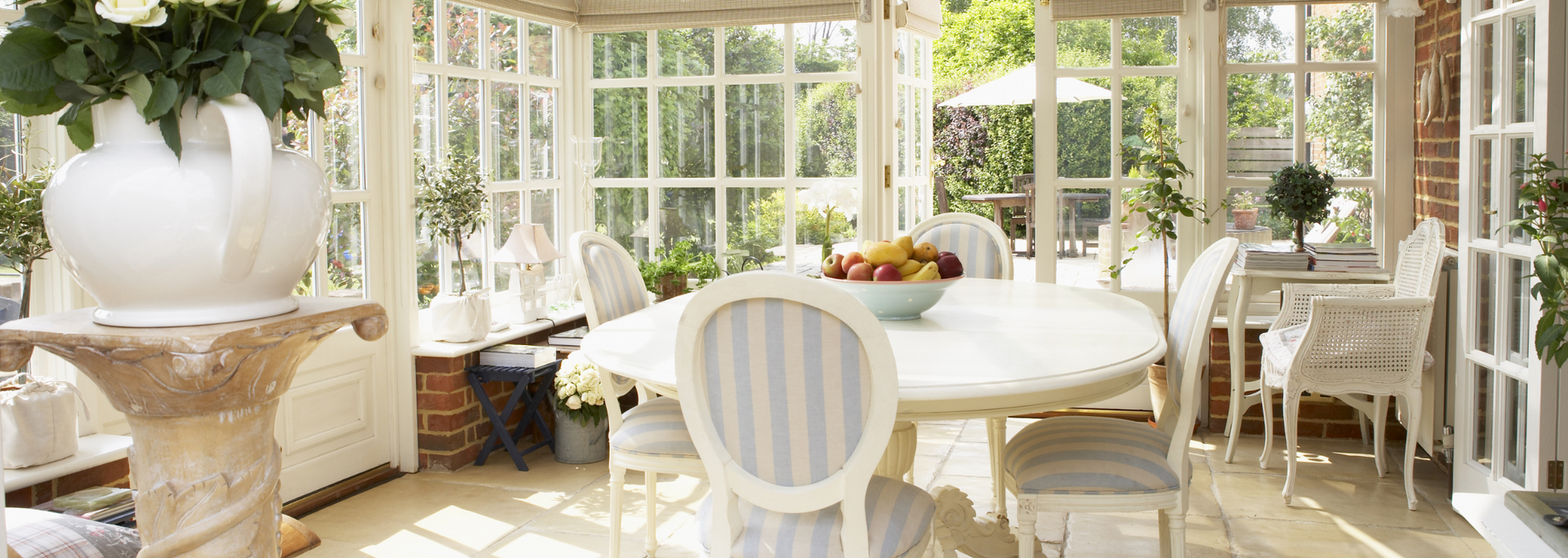 Bright sunroom with a white round table, chairs, and large windows overlooking lush greenery