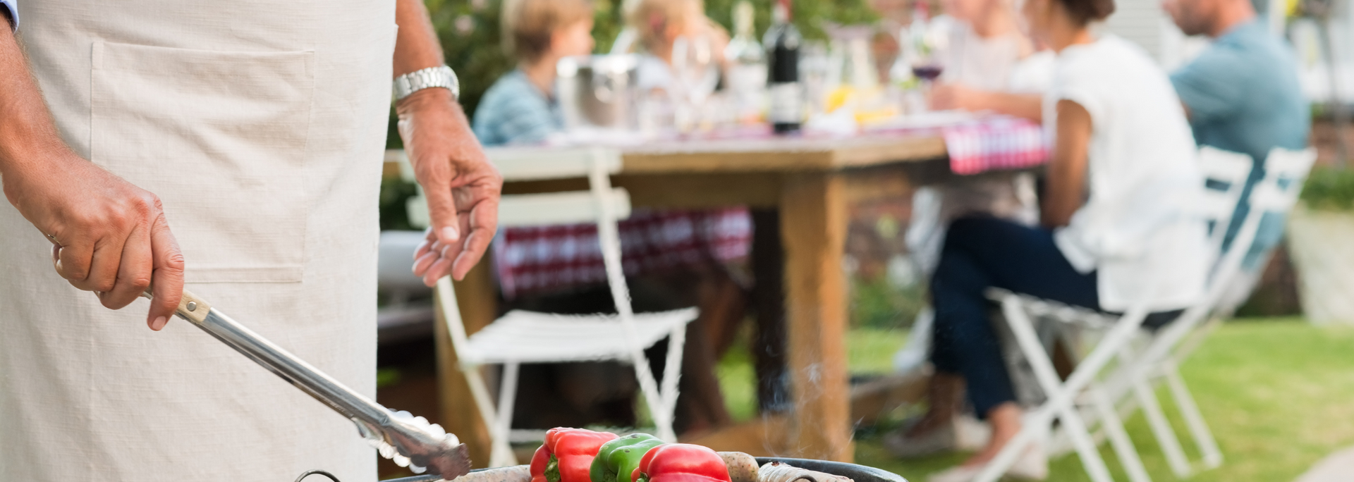 Person grilling sausages outdoors at a backyard party with a table and guests in the background