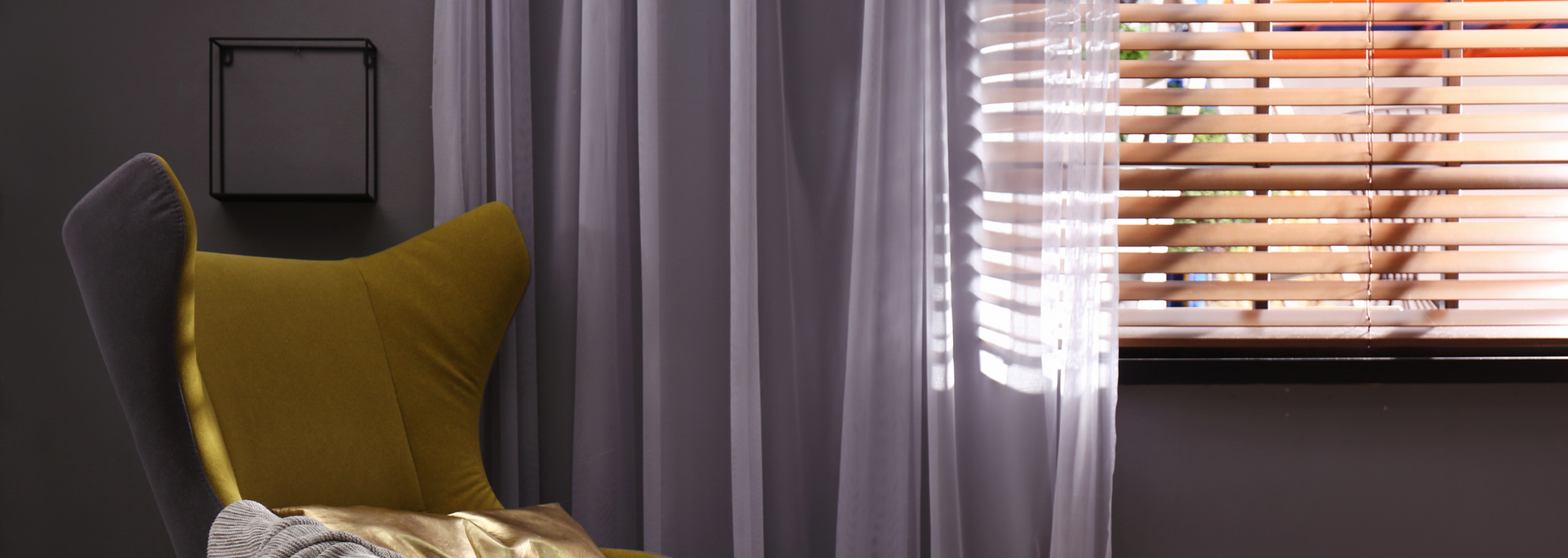 Dark bedroom corner with a mustard cushion on a chair, gray curtains, and sunlight through window blinds