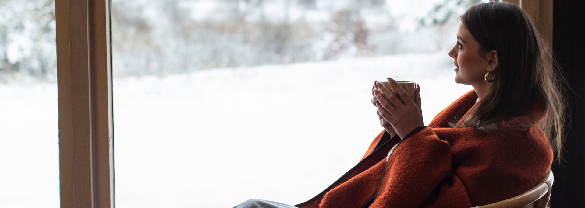 Person in a red sweater sits by a window, holding a mug and looking outside.