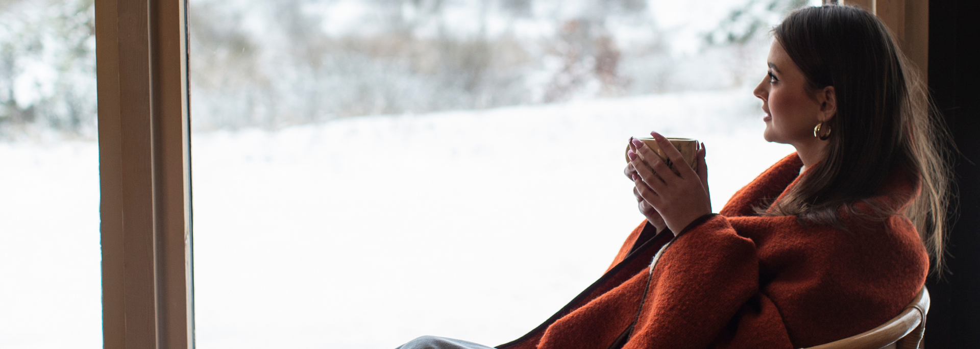 Person wrapped in a red blanket sits by a bright window, holding a cup and gazing outside.