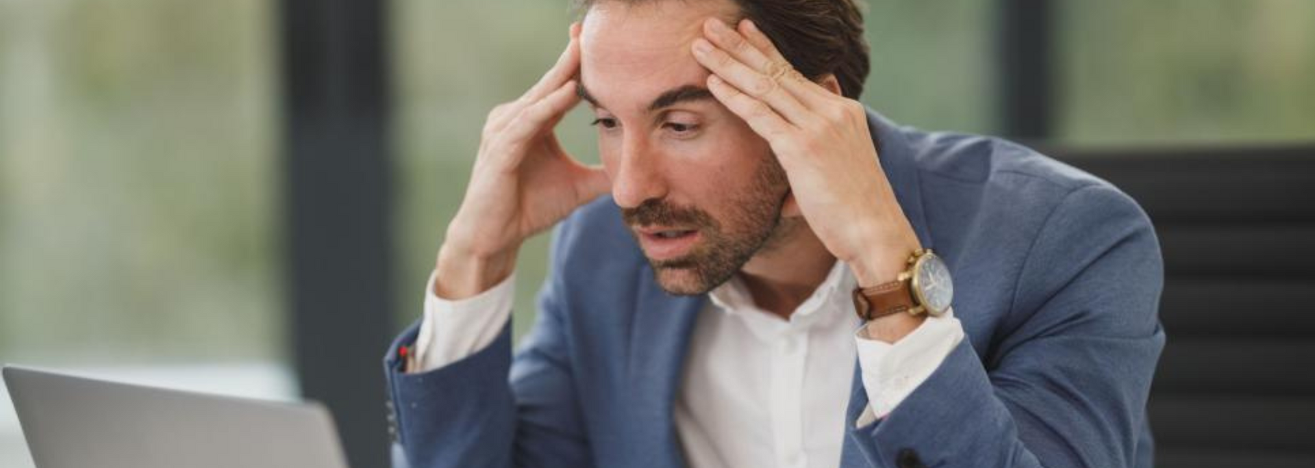 Stressed man in a blue shirt holding his head while looking at a laptop