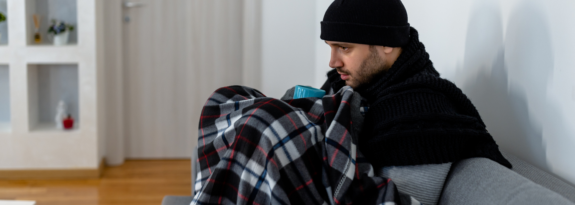 A man in a black beanie crouches beside a plaid duffel bag in a sparse room.