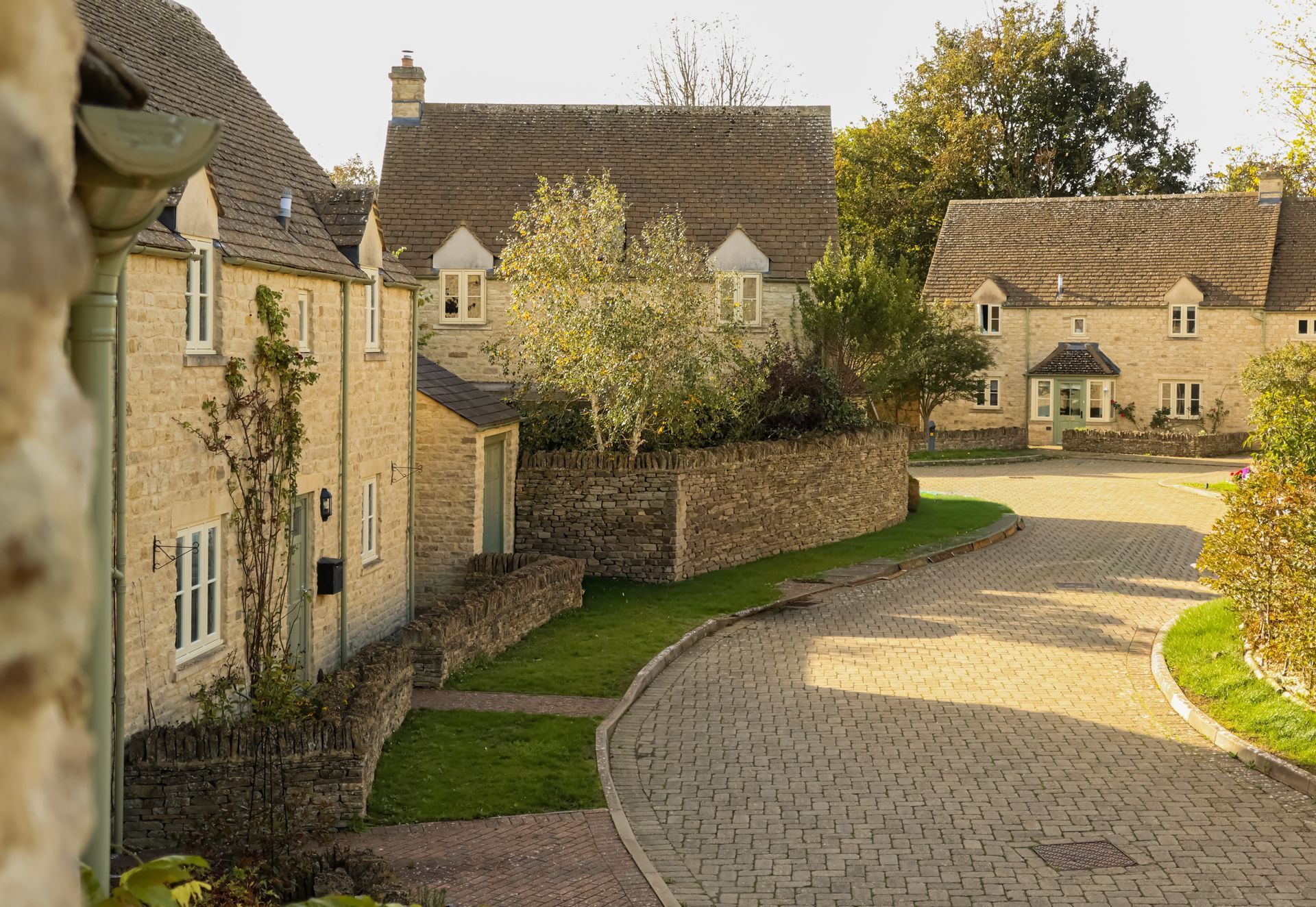 Curving cobblestone village street lined with stone cottages and green lawns in warm sunlight