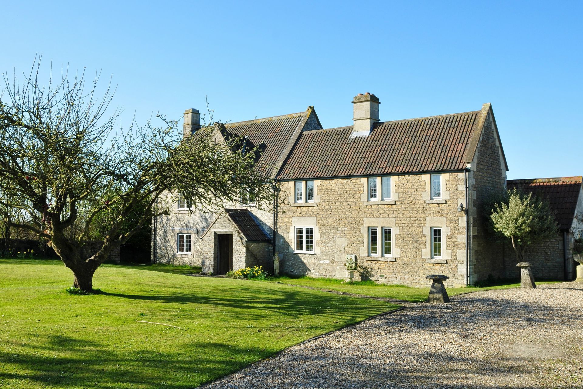 Stone countryside cottage with tiled roof, green lawn, gravel driveway, and leafless tree in front