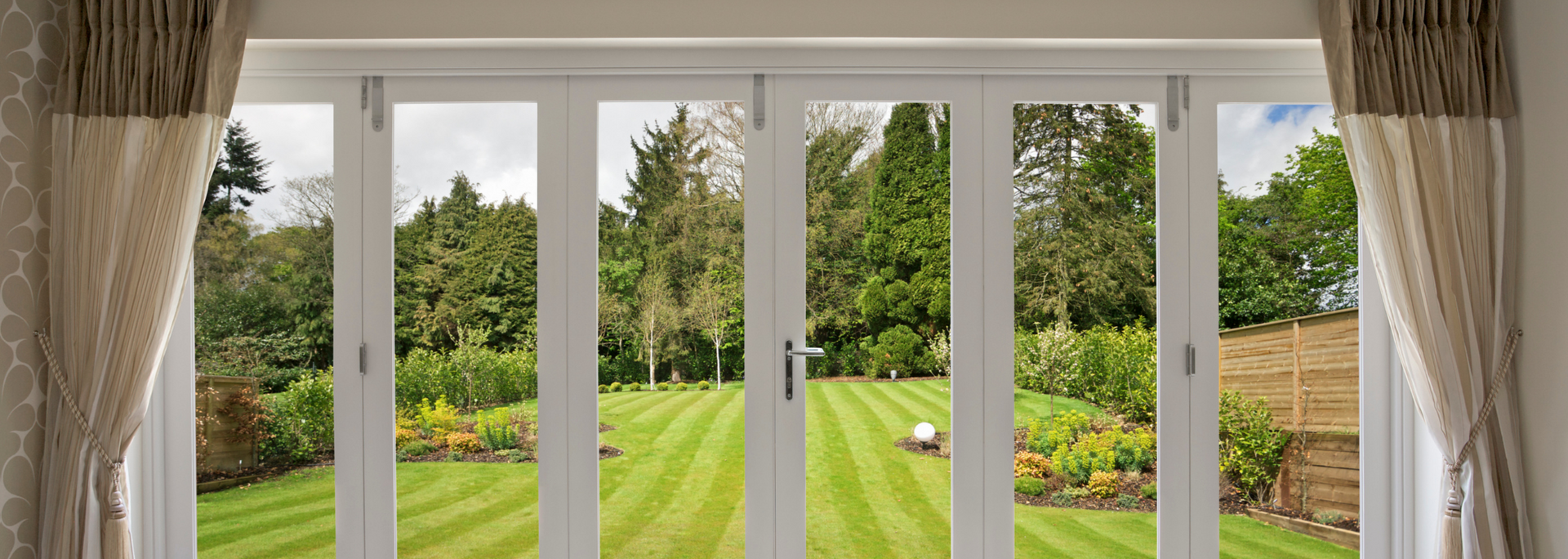 Wide garden view through white French doors and curtains onto a green lawn and trees