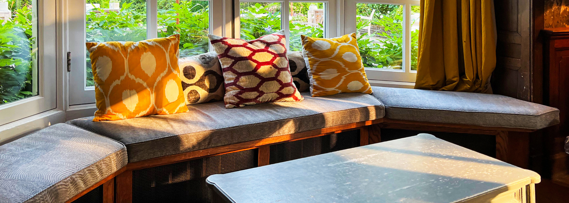 Cozy window nook with patterned cushions on a wooden bench and a small table in front of bright green garden views