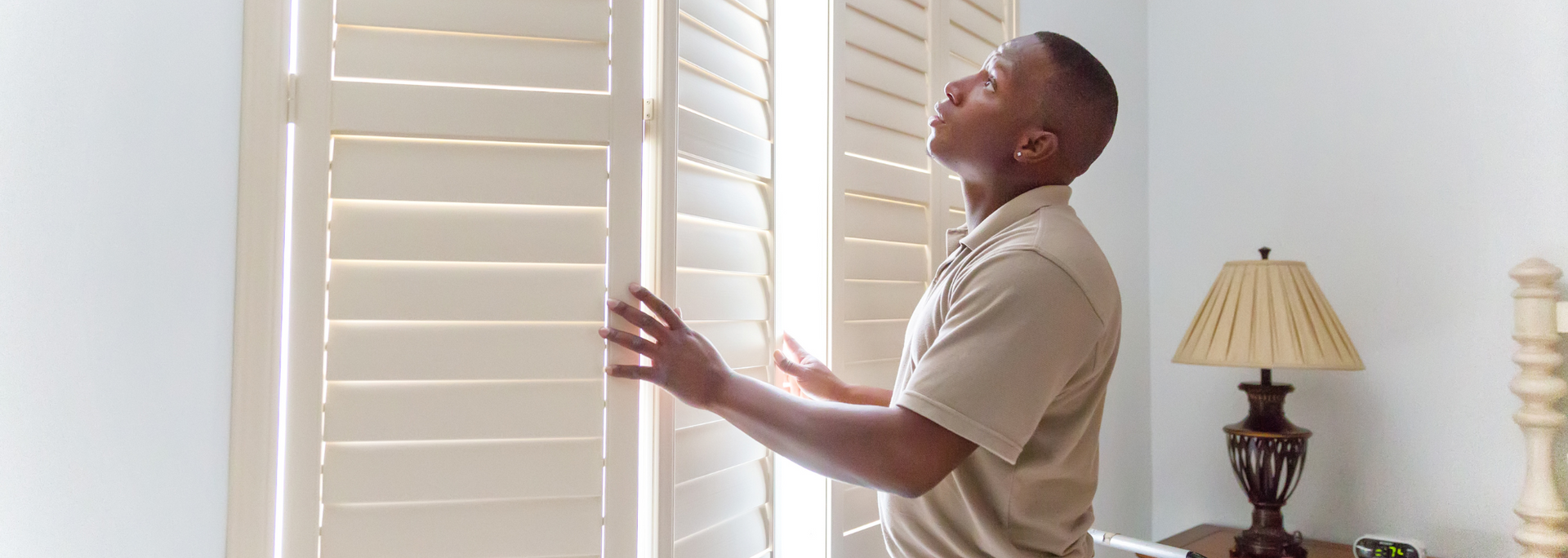 Man standing by white shutters, reaching toward sunlight in a bright room