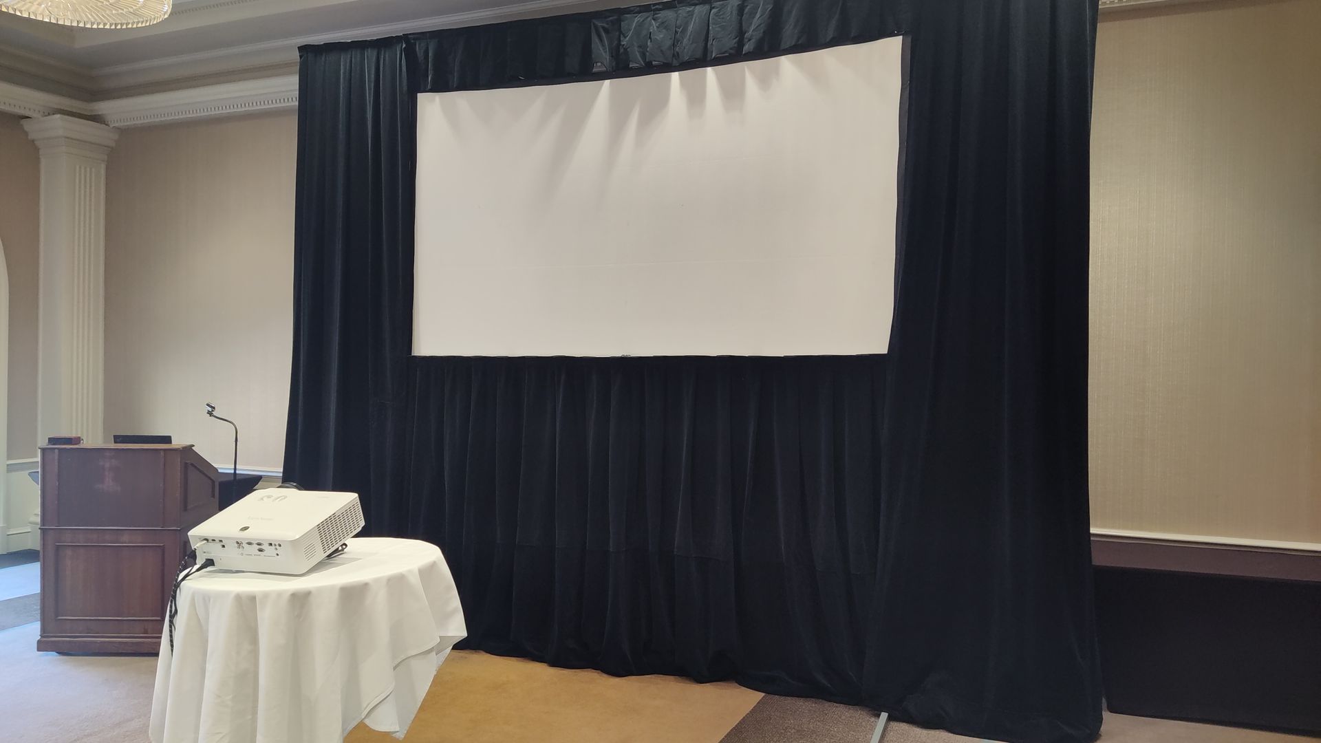 A projector is sitting on a table in front of a large screen in a conference room.