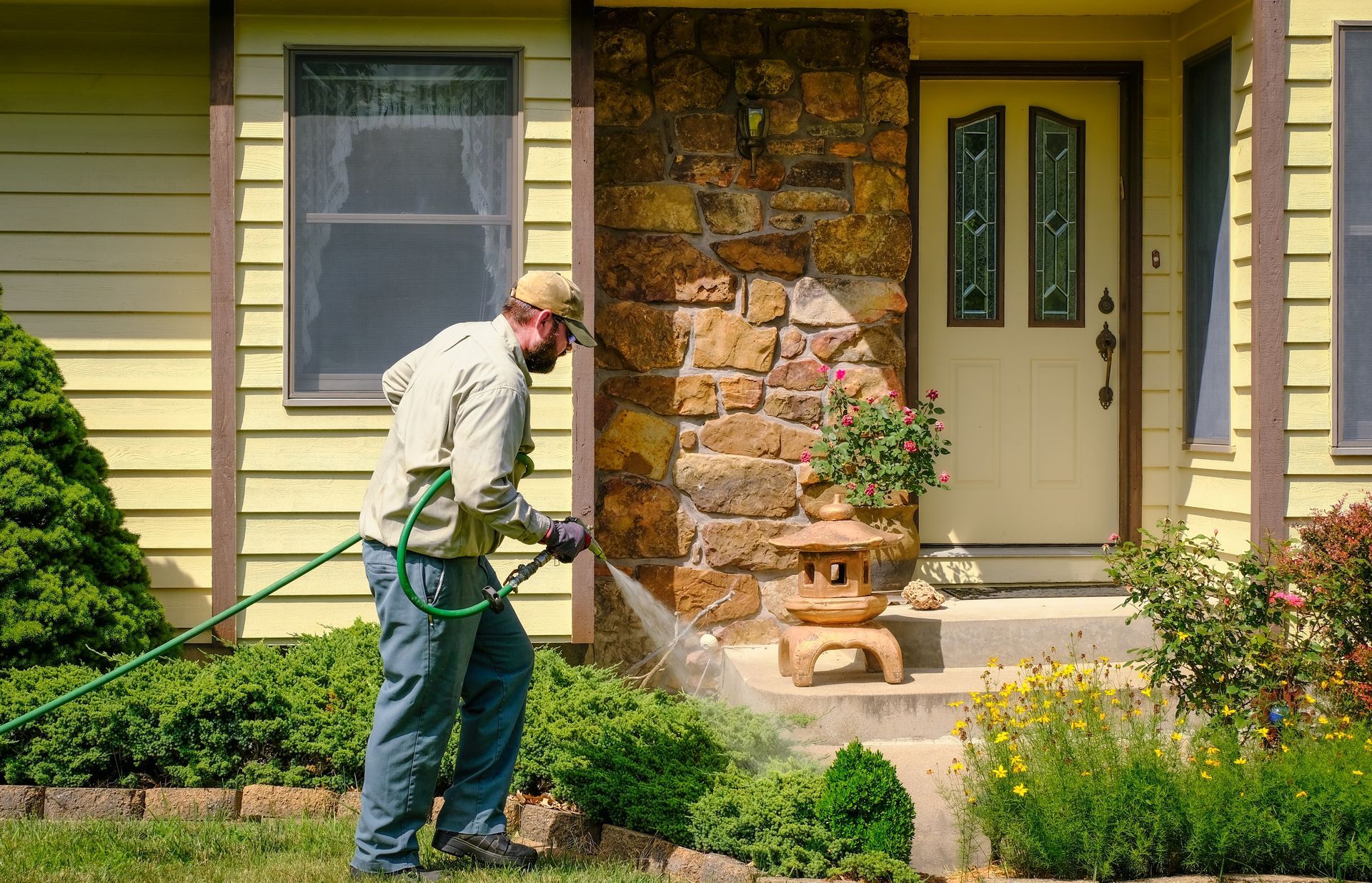 Person watering front yard plants beside a house with a hose and spray nozzle