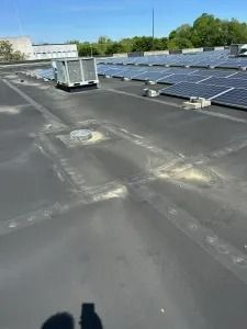 Rooftop with rows of solar panels and a large HVAC unit on a flat dark surface under a blue sky