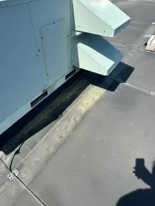 Rooftop HVAC unit beside a curb and drainage channel, with two angled vents and a shadow on the pavement
