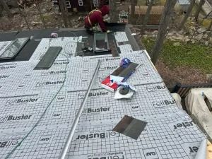 Two workers installing black roof panels with white underlayment on a sloped roof outdoors.