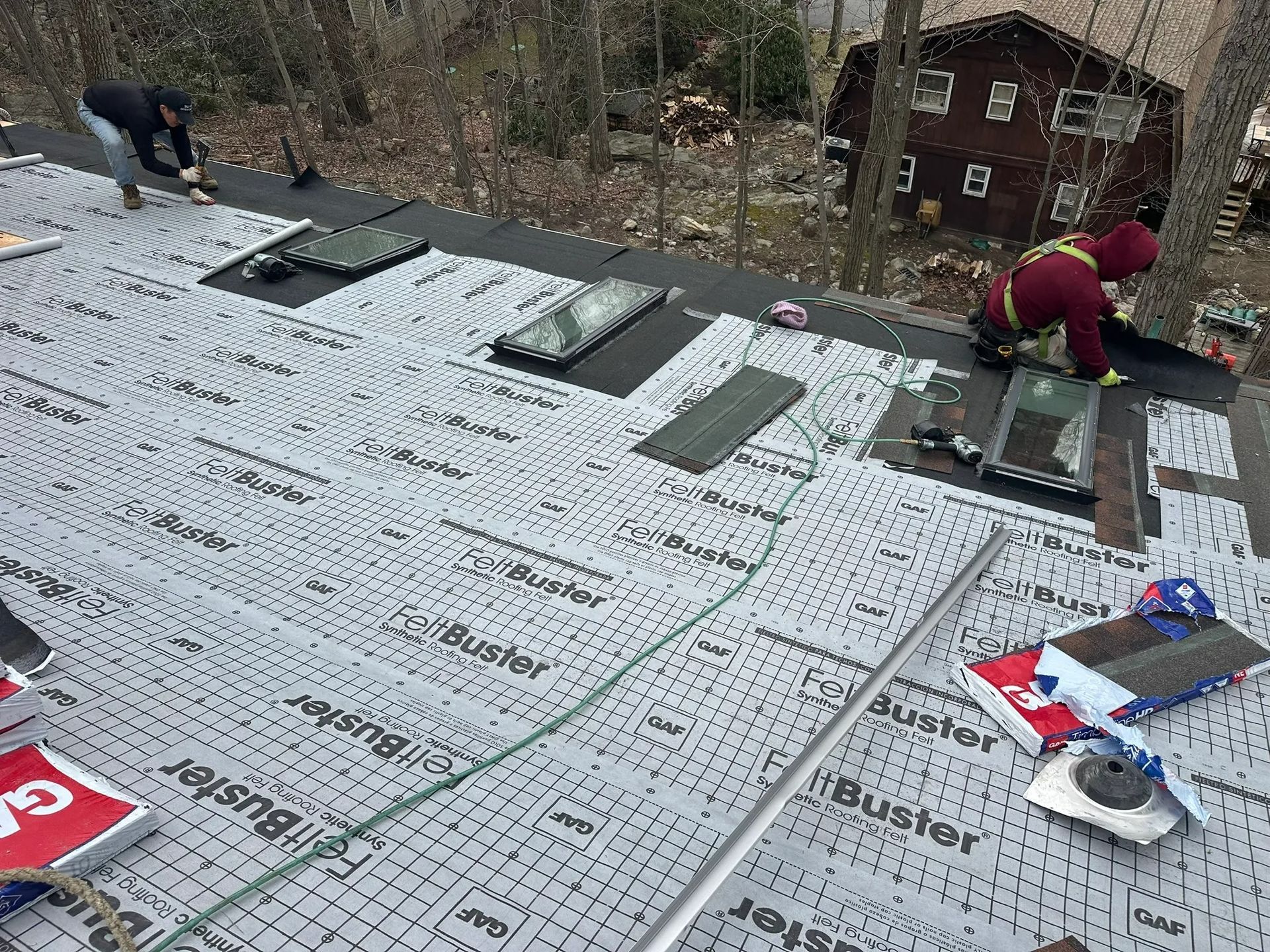 Workers installing white roofing underlayment on a roof, with tools and a wooded hillside in the background