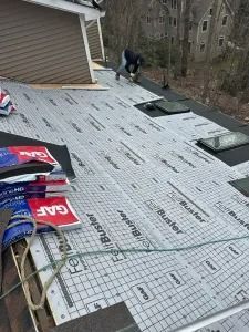 Roof underlayment being installed on a sloped roof, with a worker near the ridge and building materials in the foreground.