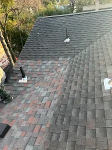 Roof with gray shingles and several white vent pipes, viewed from above beside trees.