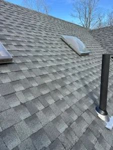 Gray shingle roof with skylights and a black vent pipe under a blue sky
