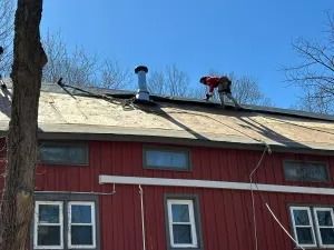 Red house with a tan roof, a chimney, and a person working on the roof near leafless trees.