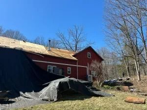 Red barn with damaged roof and tarp-covered debris in a wooded yard under a blue sky