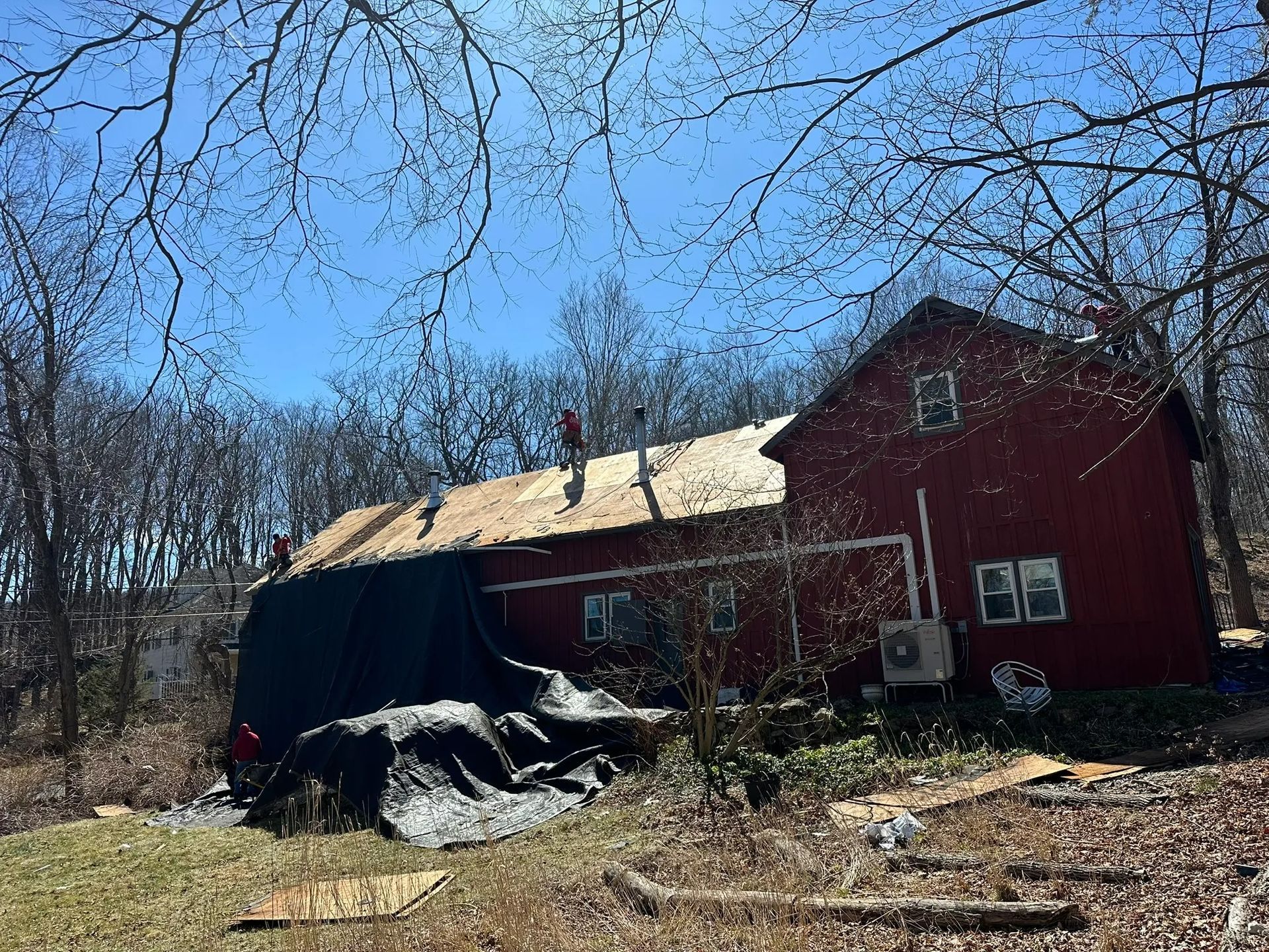 Red house with a damaged roof and debris in a wooded yard under a blue sky