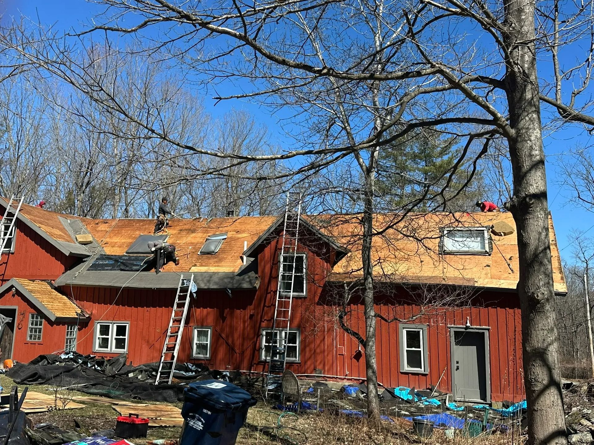 Red house with ladder on roof, several damaged windows, and a leafless yard under a blue sky.
