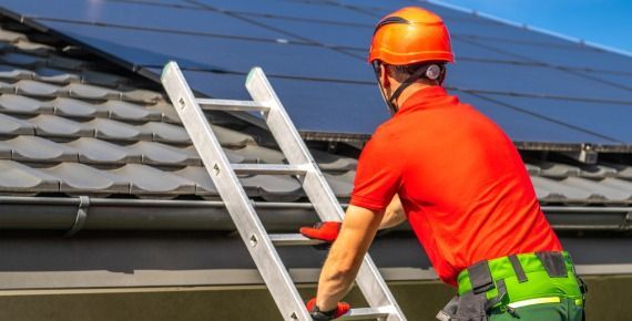 Worker in orange hard hat adjusting a ladder on a roof beside solar panels
