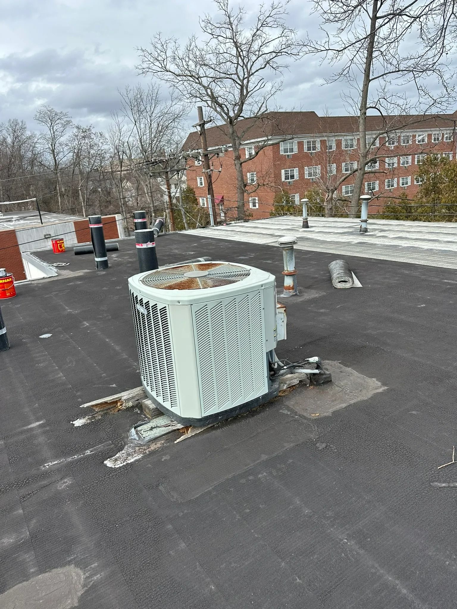 Rooftop HVAC unit on a flat roof with ductwork, snow patches, and a brick apartment building in the background