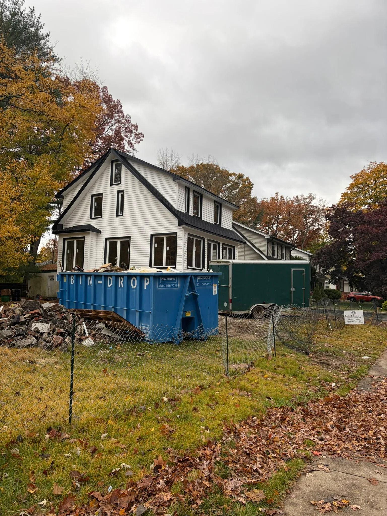 White house with blue dumpster in yard, surrounded by autumn trees under a cloudy sky