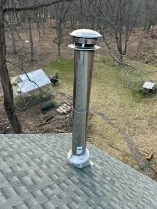 Stainless steel chimney vent on a shingled roof, overlooking a wooded yard with a small shed.