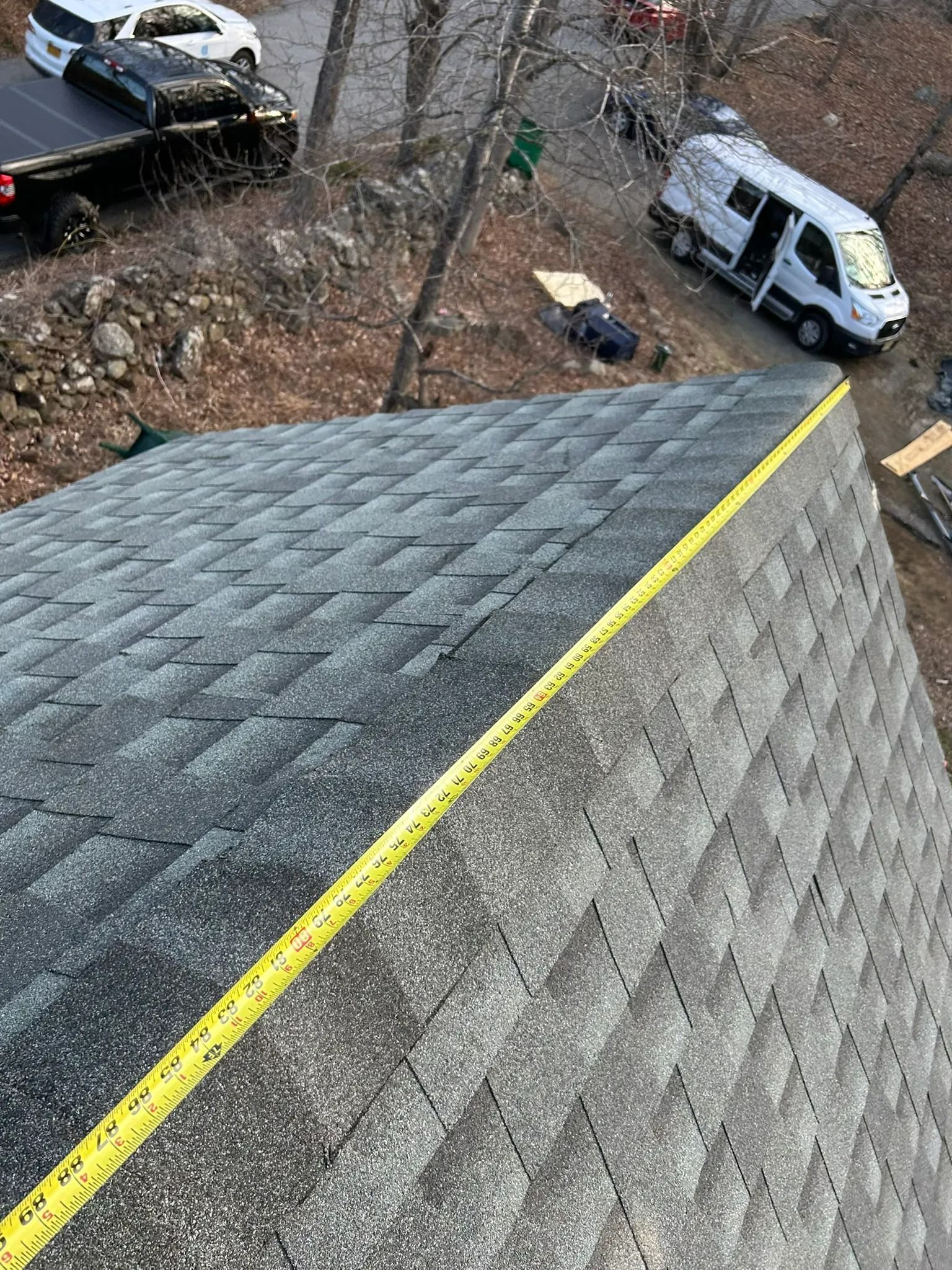 Roof shingles in the foreground with a yellow ridge line, viewed above parked vehicles and bare trees below.
