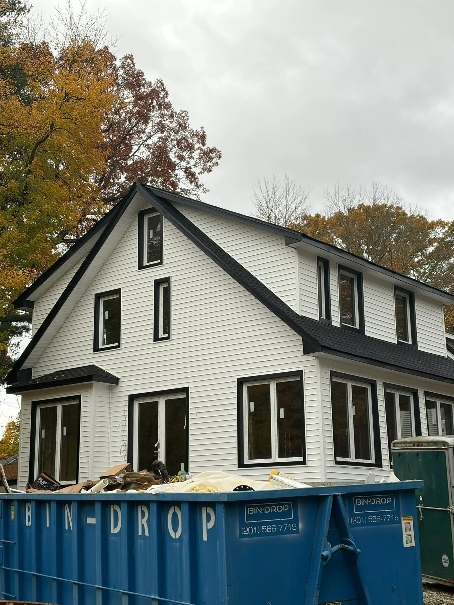 House under renovation with blue dumpster in front, white siding, and autumn trees behind