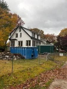 White house with black trim beside a blue dumpster and fence on an overcast autumn day
