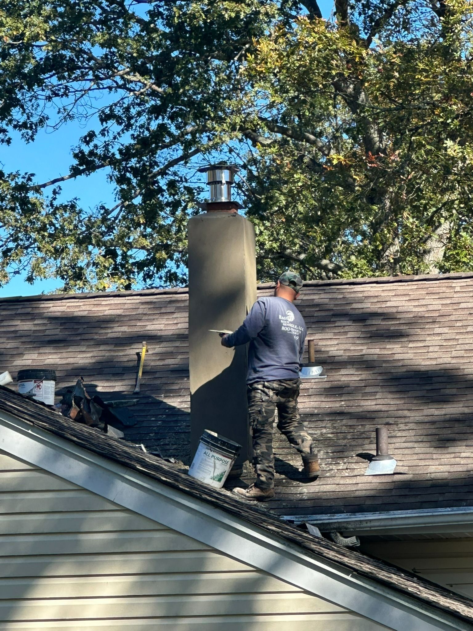Worker repairing a shingled roof beside a large chimney under a blue sky