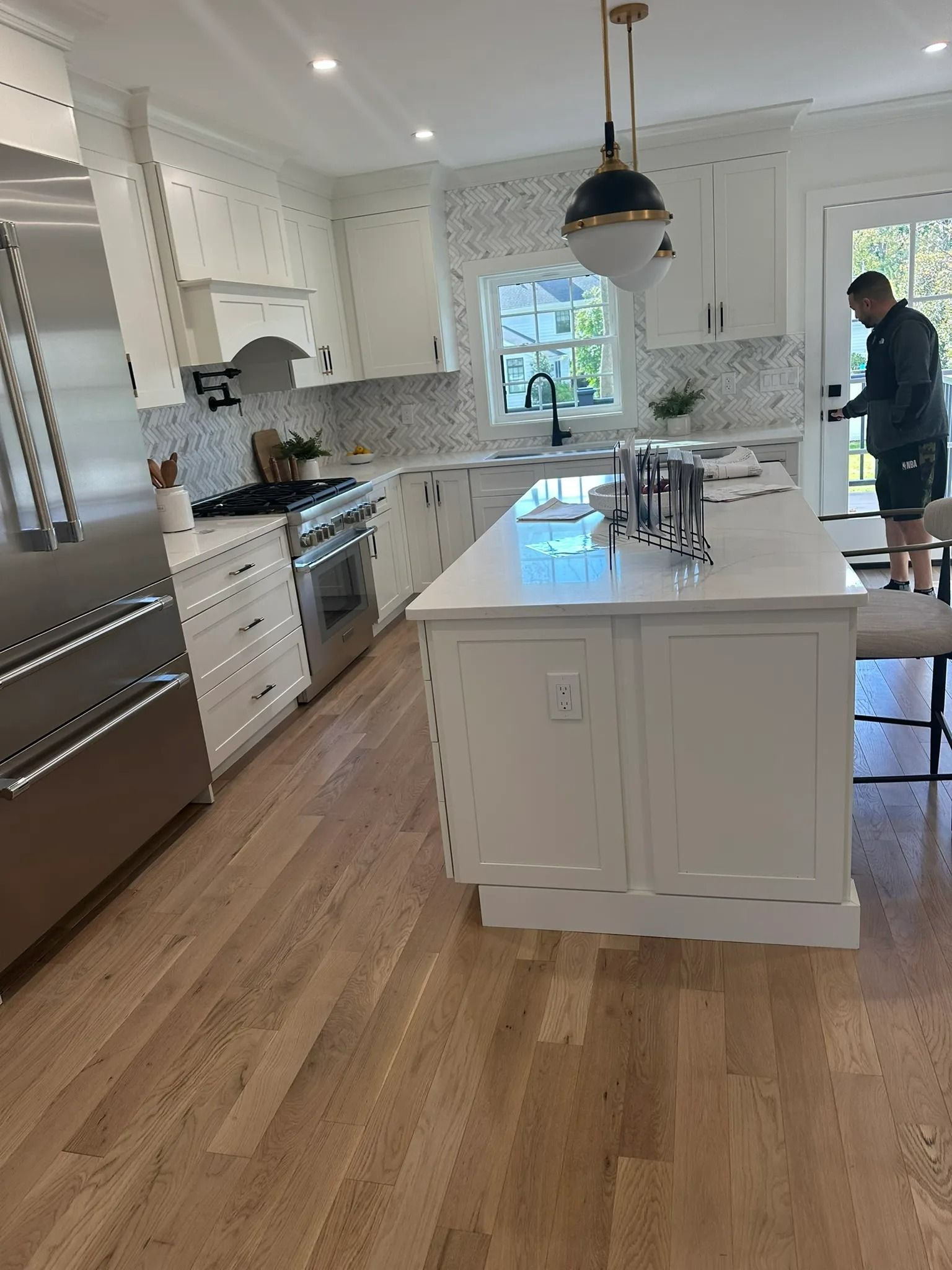 Bright white kitchen with island, stainless appliances, wood floor, and a person near the doorway.